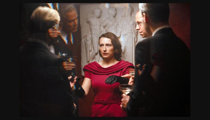 Una mujer vestida de rojo rodeada de hombres, en Woman Unknown.